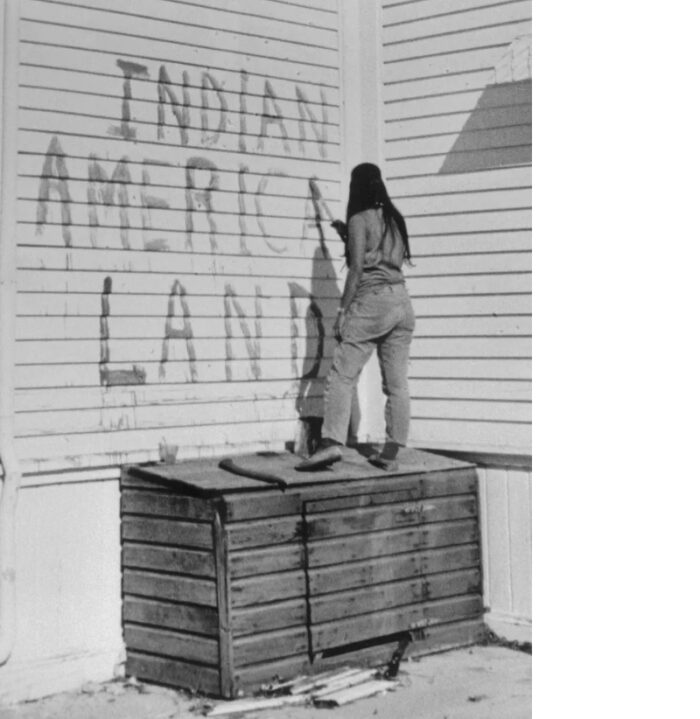 Eine Aktivistin schreibt mit einem Pinsel in der Hand in großen Buchstaben etwas an eine Hauswand: Indian America Land