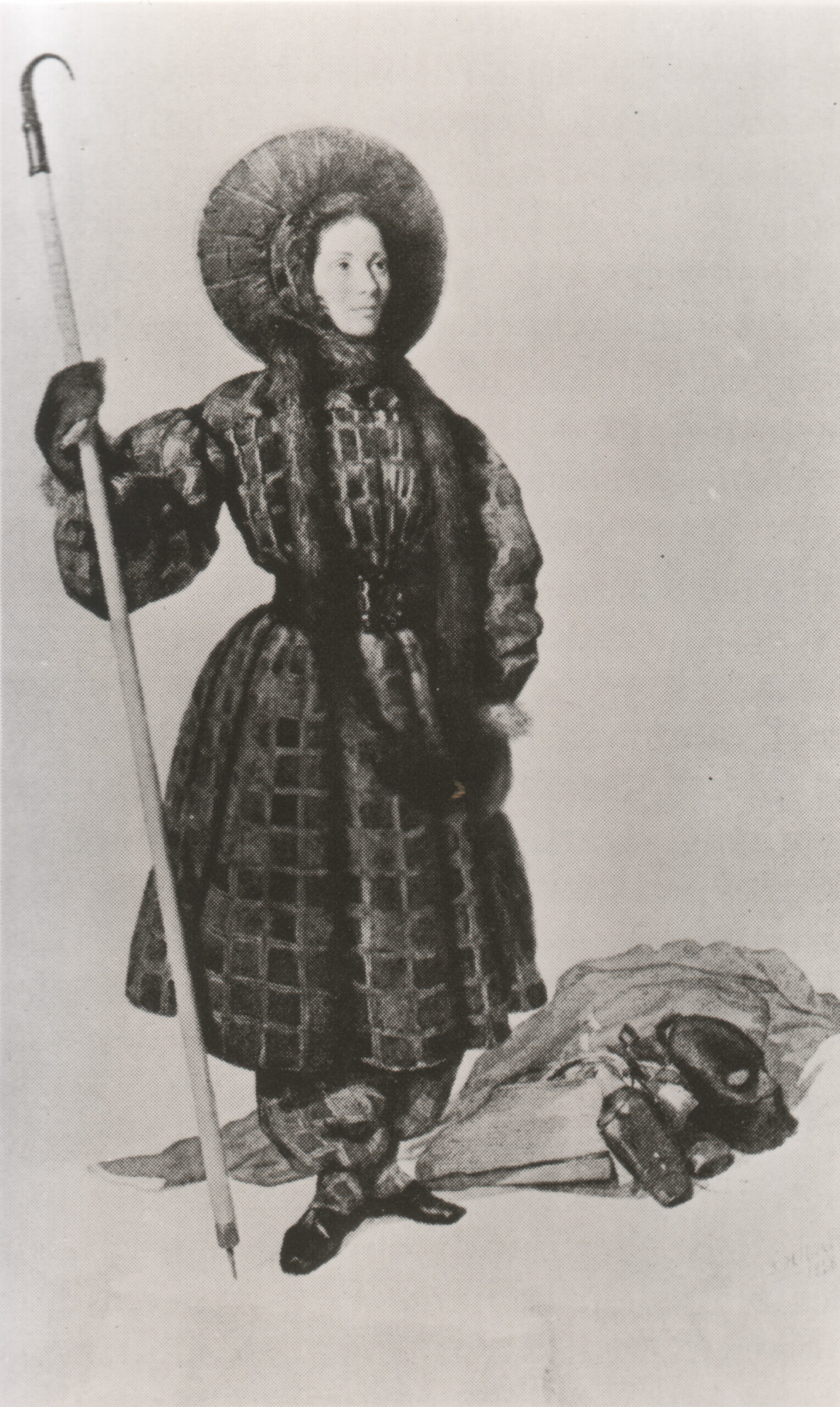 Zeichnung der Bergsteigerin Henriette d'Angeville in der Kleidung, in der sie auf den Mont-Blanc gestiegen ist. Sie trägt einen Mantel und Pluderhosen. In der Hand hält sie einen langen Stab.